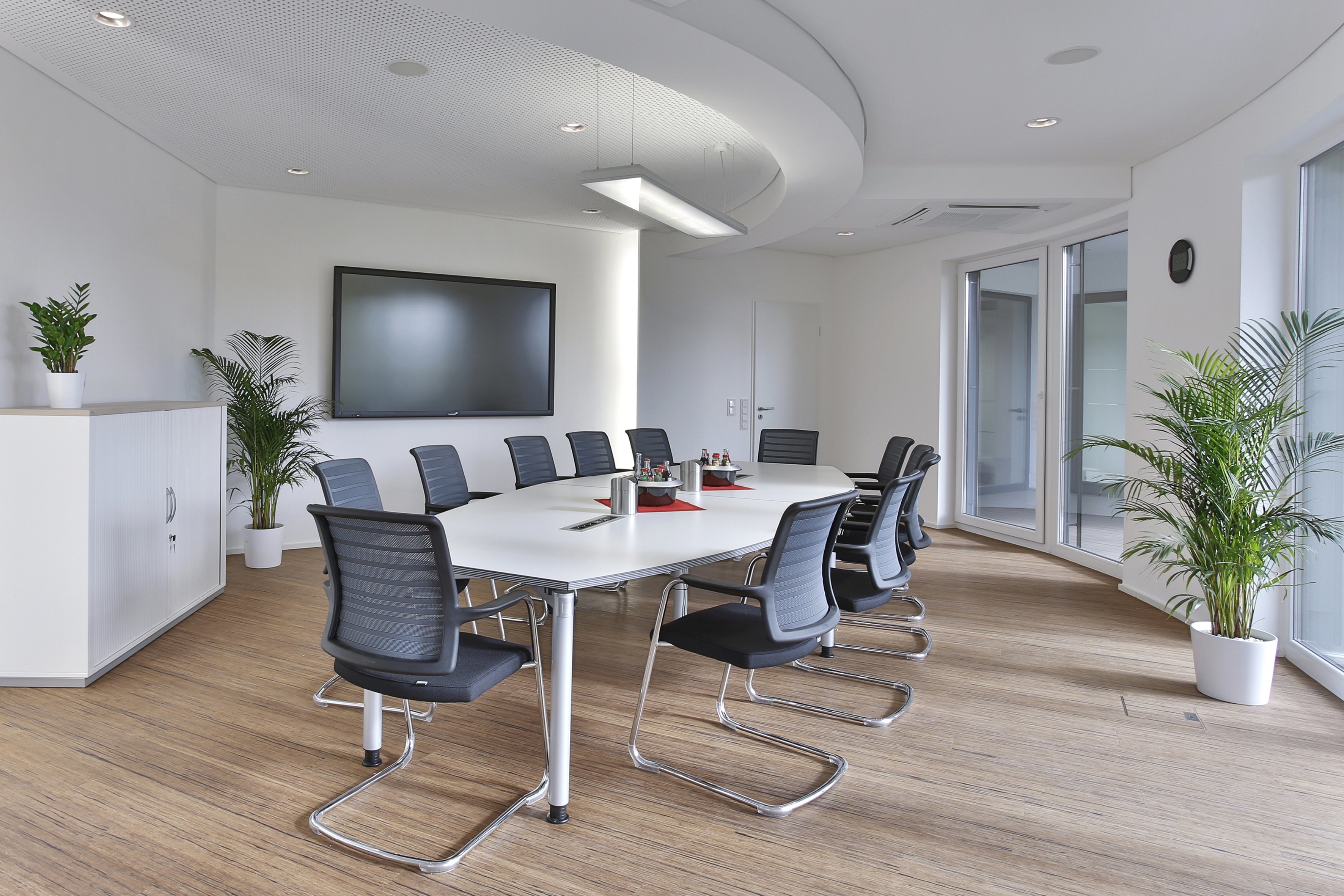 Besprechungsraum mit weißem Tisch, schwarzen Stühlen und großem Bildschirm. Palmberg Büromöbel für moderne Arbeitswelten.
