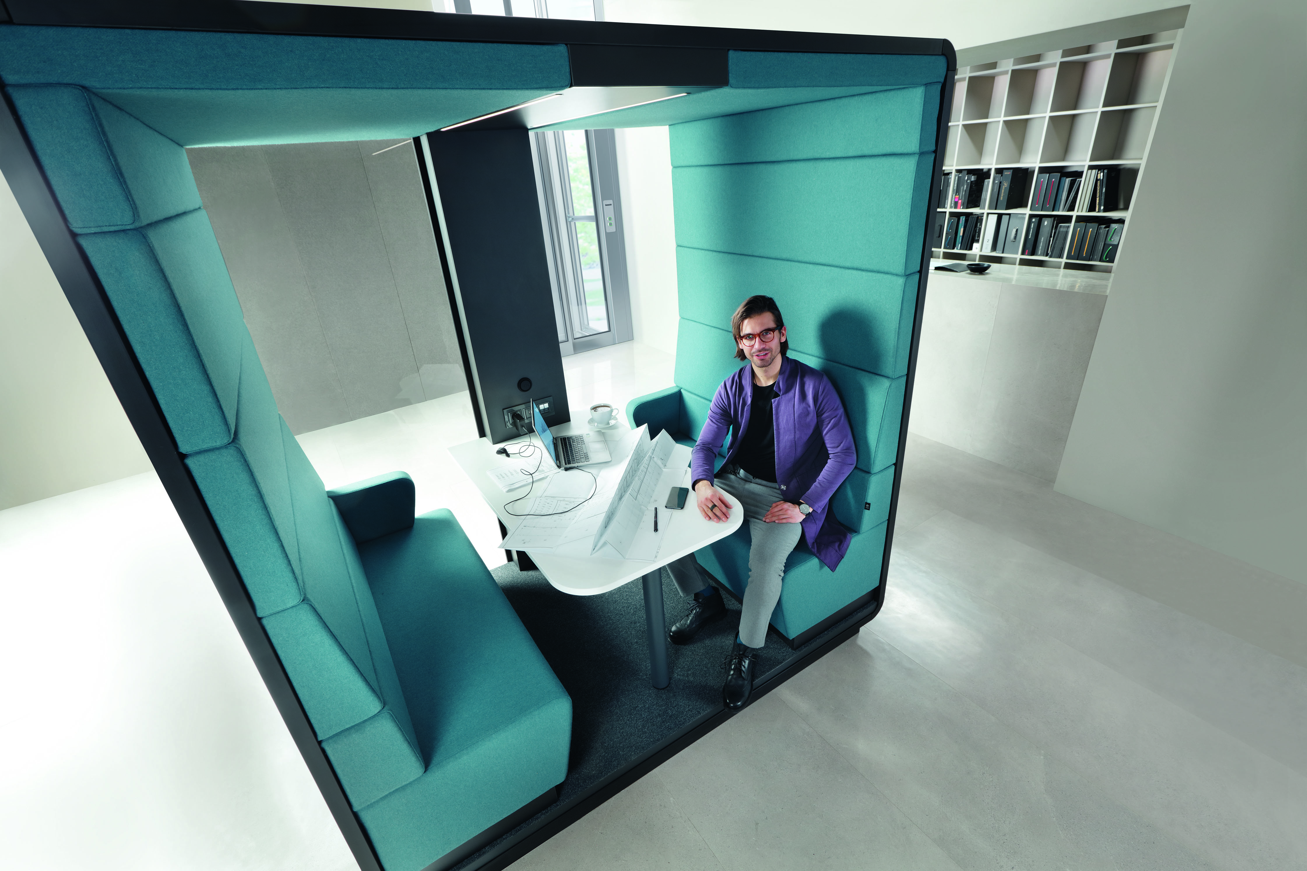 Mann sitzt in blauer Akustikkabine mit Tisch, Laptop und Bauplänen für konzentriertes Arbeiten.