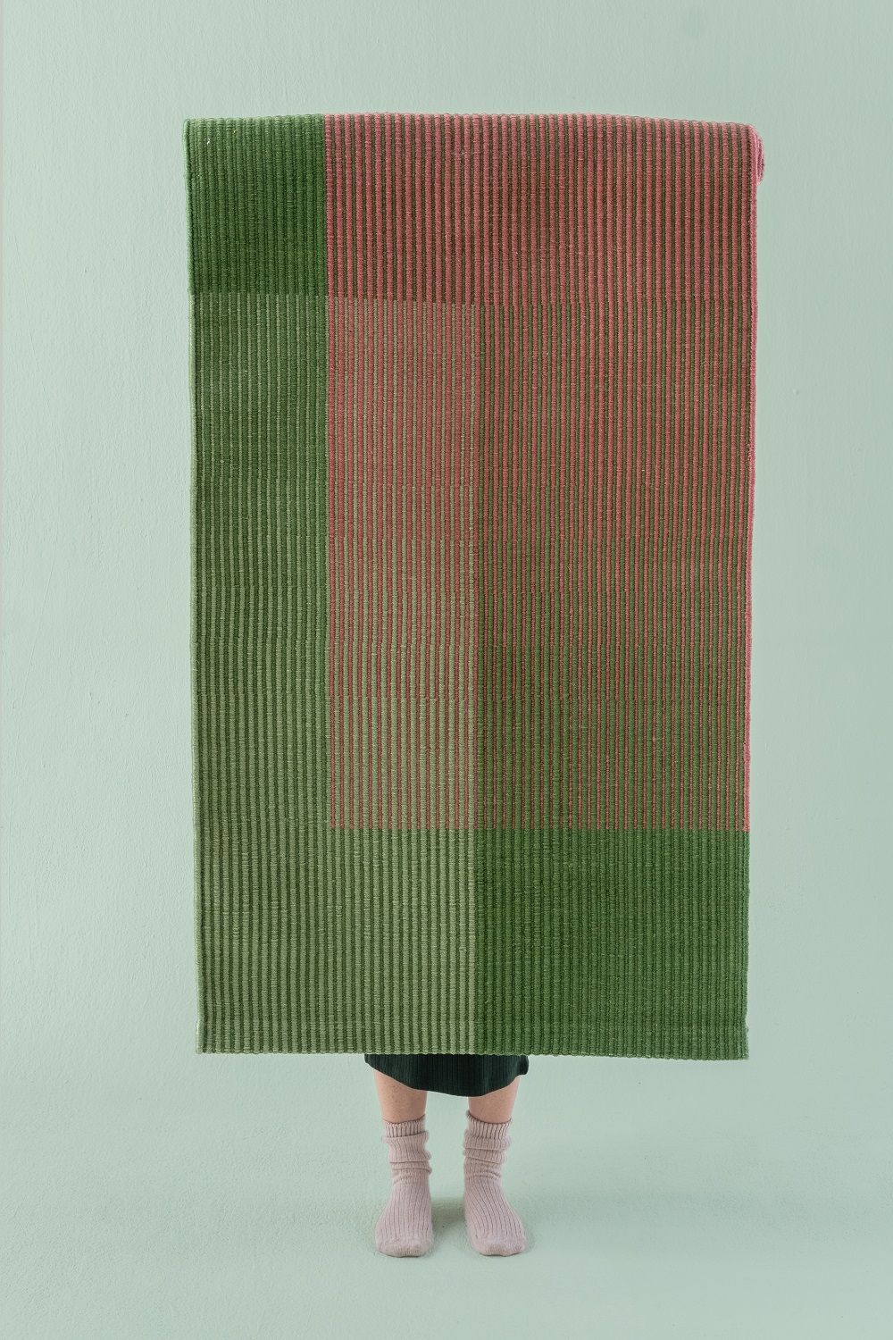 Haze Rug Teppich von Nanimarquina in Grün und Rosa, vertikal gestreift, vor hellblauem Hintergrund.