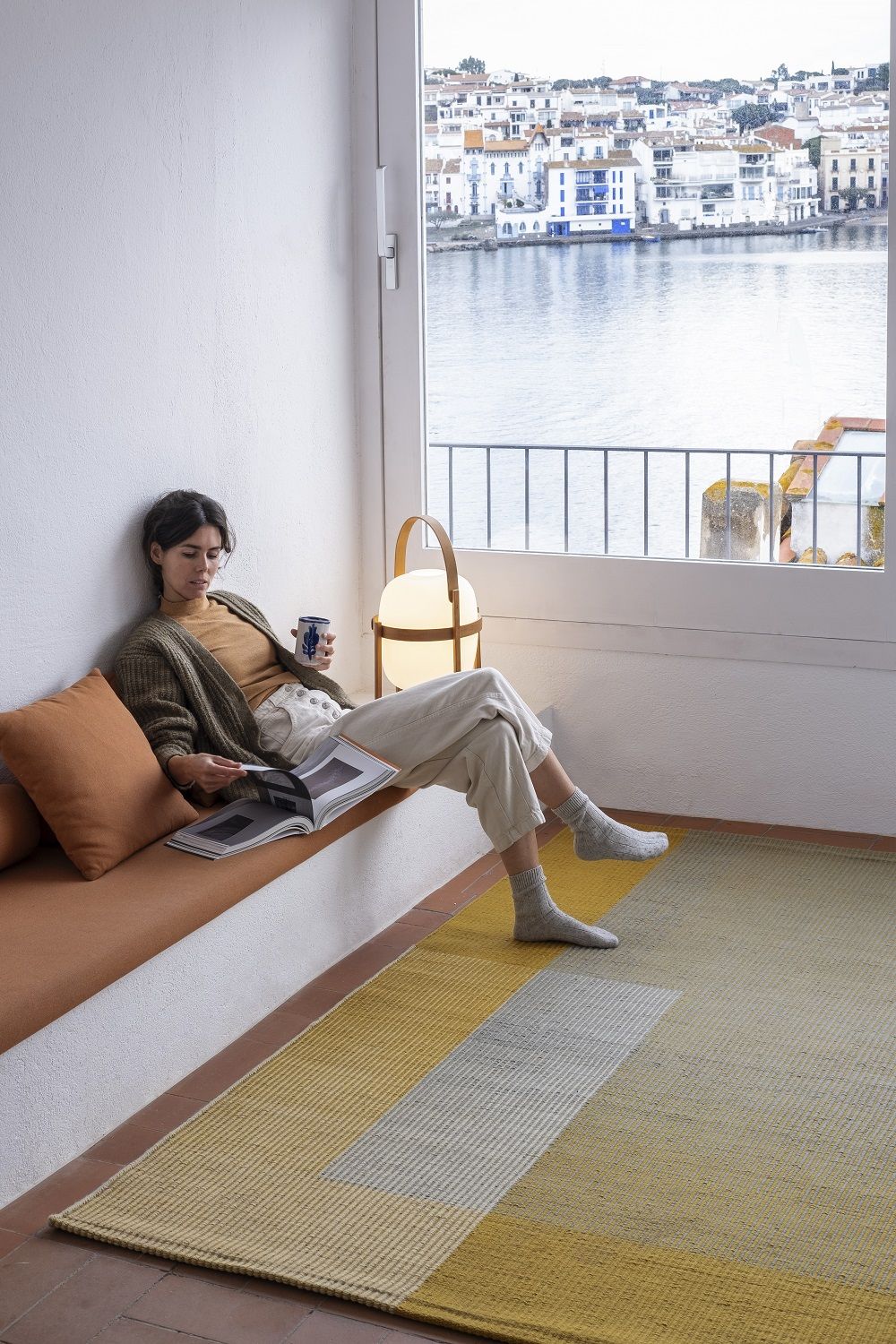 Frau entspannt auf Bank mit Buch, Nanimarquina Haze Rug Teppich in Gelb-Grau im Vordergrund.