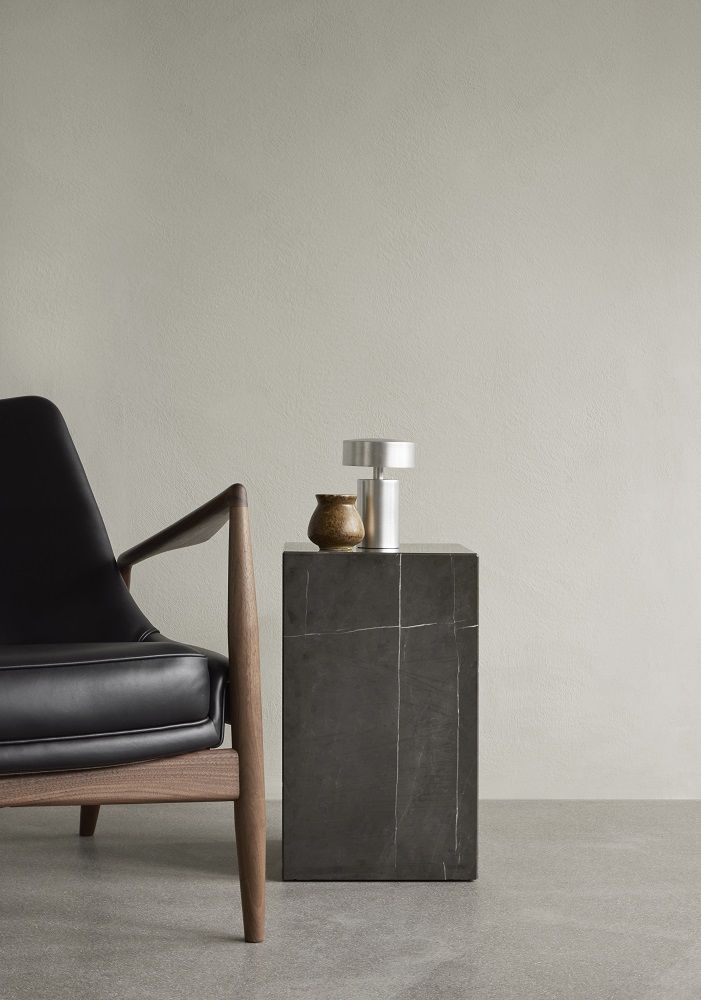 Audo Copenhagen Column Tischleuchte aus Aluminium auf einem dunklen Beistelltisch neben einem Sessel.