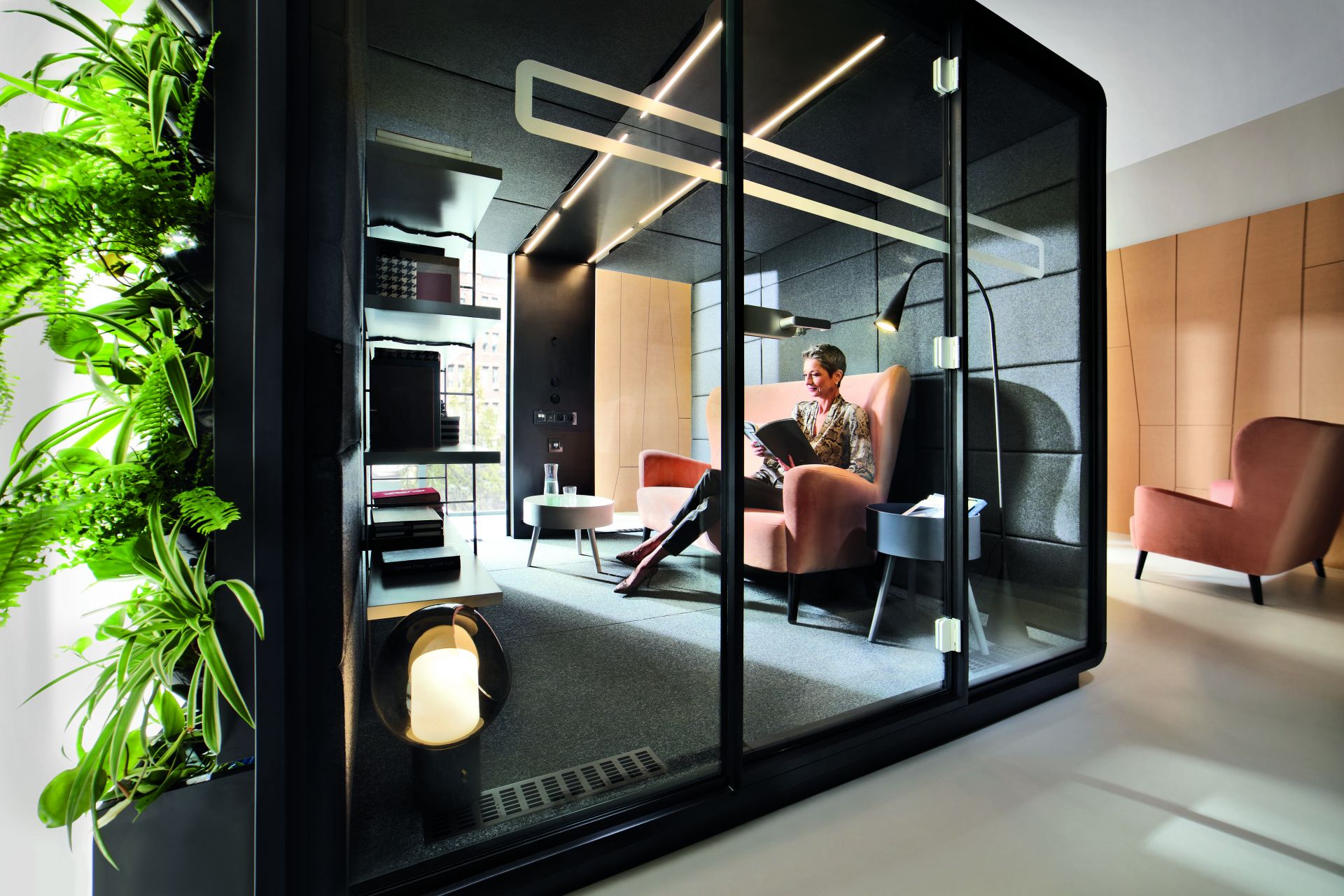 HushMeet L Meetingbox: Frau liest in moderner Telefonzelle/Besprechungsraum mit Glaswänden und Pflanzenwand.
