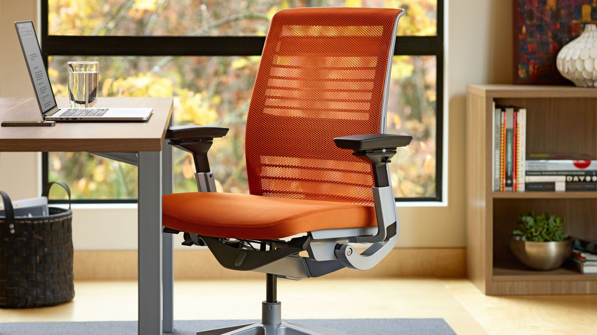 Ergonomischer Steelcase Think Bürostuhl mit orangefarbenem 3D-Rückennetz und verstellbaren Armlehnen am Schreibtisch.